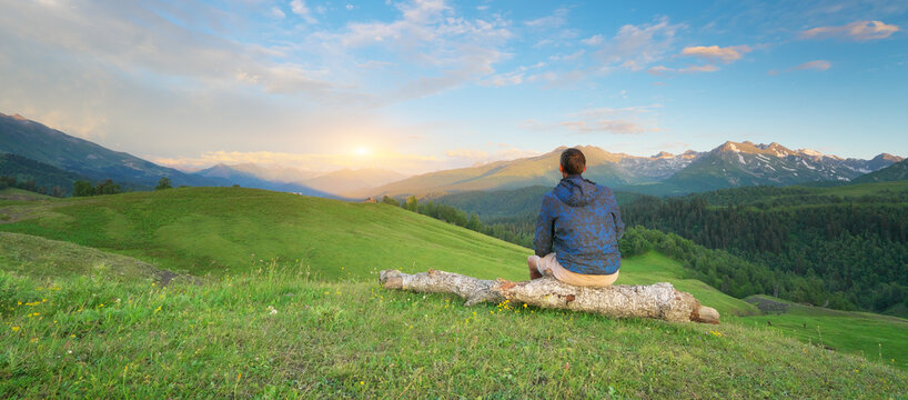 Man traveler sitting on a wooden log and looking at a beautiful sunset over mountain valley. Young tourist enjoying scenic alpine nature landscape panorama