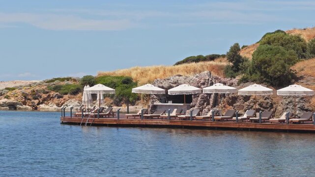 High-quality 4K wide shot of an elegant wooden sun deck over the calm sea at a luxury beach resort. Features white umbrellas, loungers, and a seaside bar.