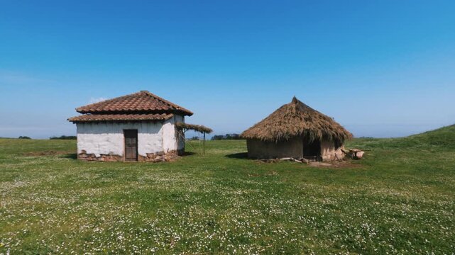 Frontal tracking shot of reconstructed ancient Astur Roman huts on a green grass field under a clear blue sky at La Campa Torres archaeological park in Gijon, Spain.