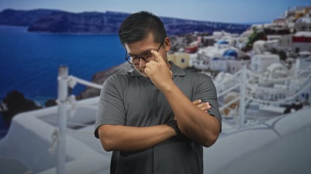 Man adjusting glasses with arms crossed on a building terrace overlooking blue sea and white domed houses; contemplation.