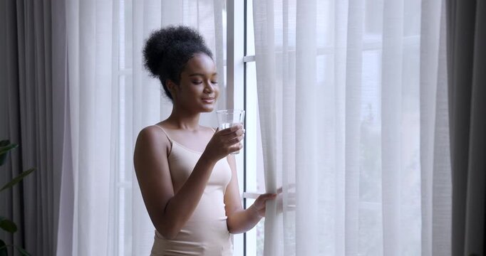 multiracial woman holding glass water standing near window looking outside feeling relaxed hydrated skin glowing slowmotion light passing through curtain enhancing natural beauty in calm morning