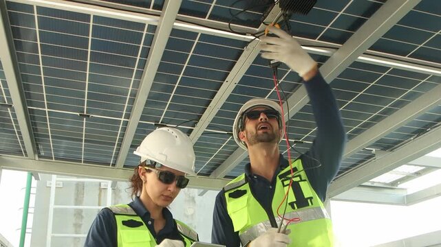 Engineers with safety vest and helmet work with electrical instrument on photovoltaic solar panels together. Male technician uses multimeters to check solar power, woman worker enter data into tablet