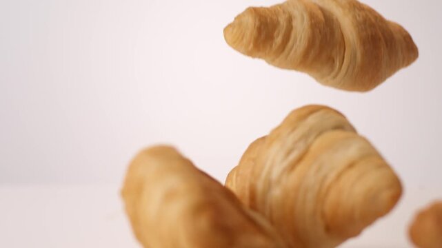 Golden buttery flaky croissants bouncing and tumbling on white in slow motion macro studio shot. french bakery branding, parisian cafe content, brunch menu reels.