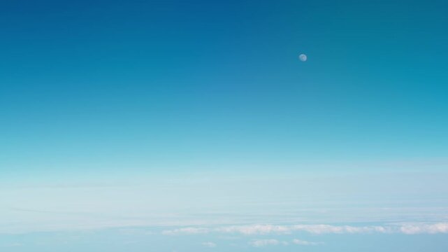 Daytime moon seen on an airplane passenger window and blue sky over clouds airborne, handheld