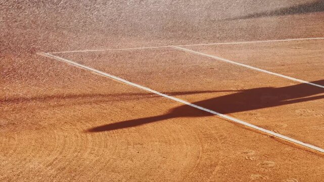Clay tennis court being watered in warm sunlight, with white boundary lines, wet orange surface, long shadow, and sports venue maintenance detail.