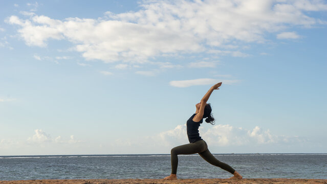 Woman Practicing Warrior Yoga Pose on Ocean Coast Under Blue Sky