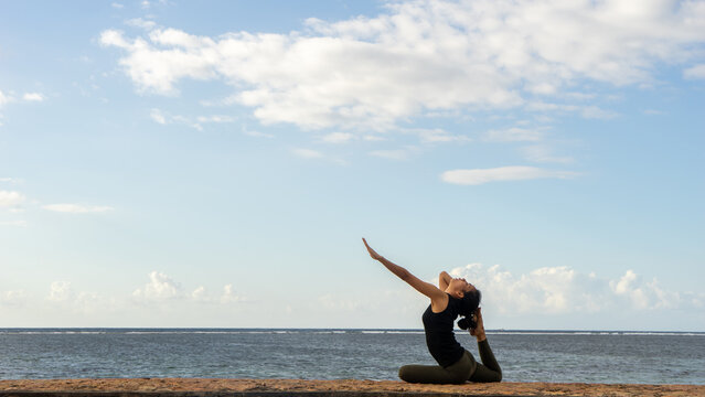 Woman Practicing King Pigeon Yoga Pose on Ocean Coast Under Blue Sky
