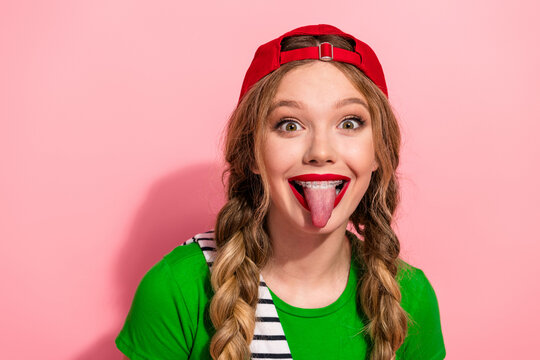Playful young model with braids wearing a green shirt red cap sticking out her tongue against pink background