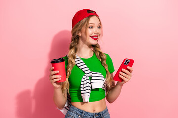 Obraz premium Young stylish girl with green top red cap holding coffee and phone against pink background looking happy and fashionable