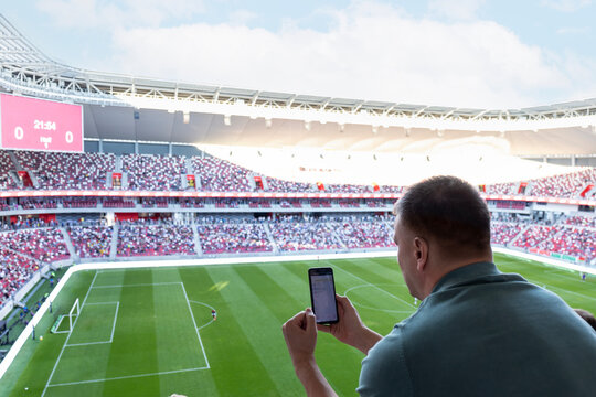 Caucasian man holding smartphone with sports bets app while watching football in the stadium