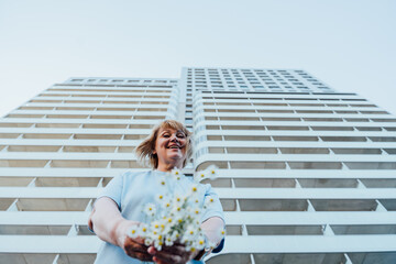 Smiling Senior Woman Holding Daisy Bouquet Near Modern Residential Tower © Iryna