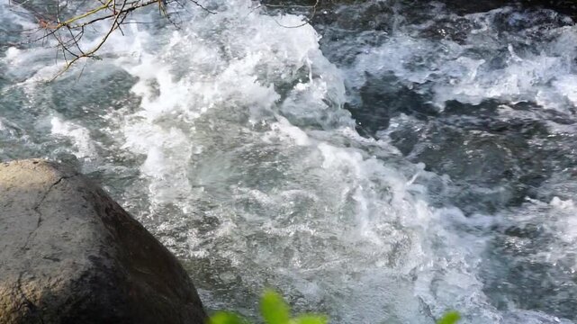 Mid-shot video in 4x slow motion turbulent mountain river with crystal-clear water tumbling over rapid, next to exposed rocks and boulders. Angle is from side-on view, overhead perspective, 30 fps