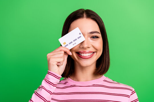 Young chic woman holding a credit card smiling against a green background wearing a striped pink shirt