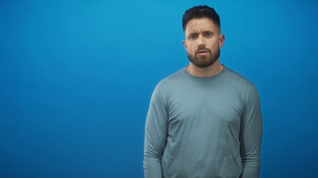 Man with beard and visible hands shrugs with palms up and raised eyebrows against a seamless blue studio backdrop; uncertainty.
