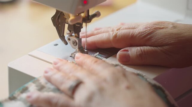 Wrinkled hands guide knit fabric through a running sewing machine and cut the thread.