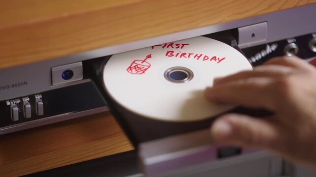 A hand slides a handwritten "First Birthday" disc into a vintage DVD player to relive the memory