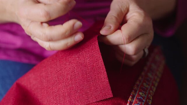 Aged hands fold and hand-stitch a red fabric hem with red thread, filmed face-on