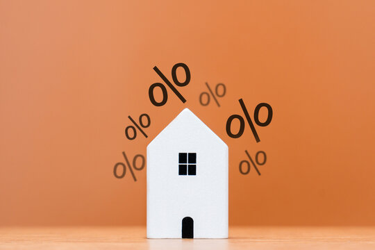 A white house model surrounded by floating percentage signs. Mortgage interest rates, property taxes, real estate investment returns, and housing market inflation concept.