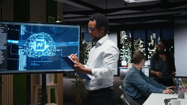Manager in office writing on notepad, taking notes of artificial intelligence system bugs to fix. Supervisor looking at large screen, documenting neural network errors during troubleshooting.