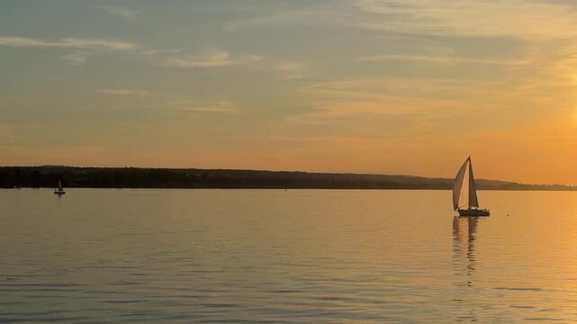 Single sailboat with white sail on calm lake at golden hour with soft reflection and distant shoreline under clear evening sky creating peaceful minimalist seascape