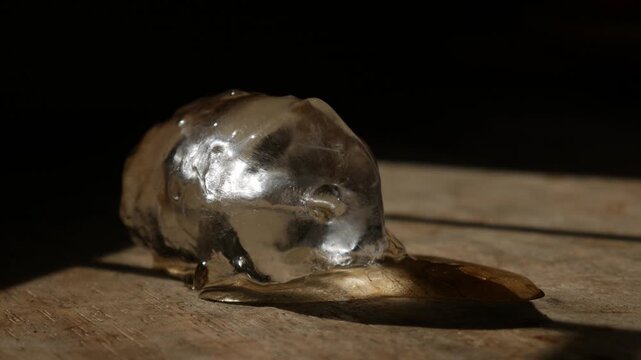 Single melting ice cube on rustic wooden table surface with dramatic sunlight shadow in dark moody still life macro photography