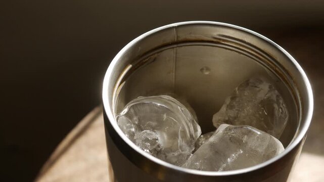 Close-up top angle view into stainless steel tumbler cup with melting ice cubes in golden warm light on wooden table in cafe