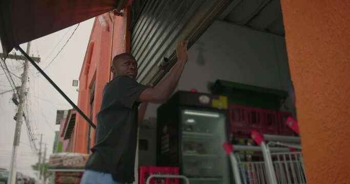 African American man closing metal storefront gate of grocery shop at end of business day on urban street with carts and products visible inside