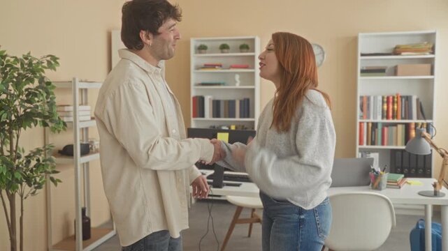 Man and woman standing face to face, man pointing while speaking and woman with clasped hands by a desk with computer, bookshelf and potted plant in a studio; tension negotiation.