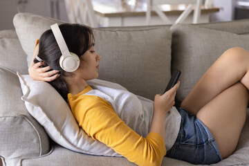 Fototapeta premium Asian woman reclining on light grey sofa using phone while wearing headphones near table and chairs