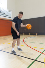 Naklejka premium Male basketball player is dribbling orange ball on hardwood court near red wall panels