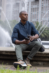 Obraz premium African man in 30s sitting on fountain ledge wearing smart-casual outfit holding phone and coffee