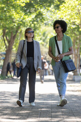 Two adults walking and talking on dappled tree-lined path, carrying crossbody bags and coffee cup © wavebreak3