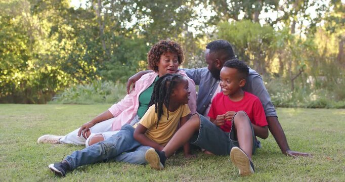 Family of four sitting on grass, mom in ripped jeans, kids talking, boy fiddling cord, bonding