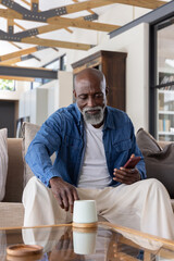 Senior African American man sitting at home in denim shirt holding smartphone and ceramic cup © wavebreak3