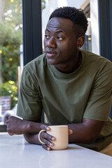 African American man sitting at countertop in olive tee holding beige mug gazing left toward glass © wavebreak3