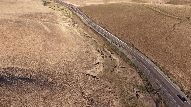 High-angle drone perspective following vehicles driving along a scenic, curving road through the dry, golden hills of the Altamont Pass in Livermore, California, on a sunny day