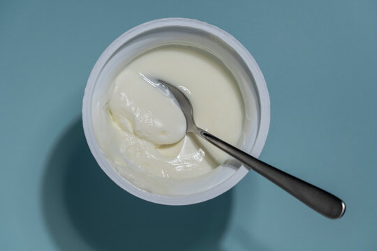 Fresh greek yogurt in a plastic cup with a spoon on blue background, closeup. Top view of creamy white yogurt or sour cream in a container with a metal spoon on a blue background