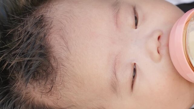 Tiny face rests softly, eyes closed. Pink bottle holds warm milk, gentle sip. Curly dark hair frames sweet, calm face. Sleeping baby drinks peacefully, no fuss. Ideal for parenting, baby care