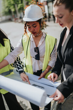 Female construction professionals wearing hard hats and safety vests examine rolled blueprints on site. A team of engineers and architects reviews plans together during an outdoor project meeting.