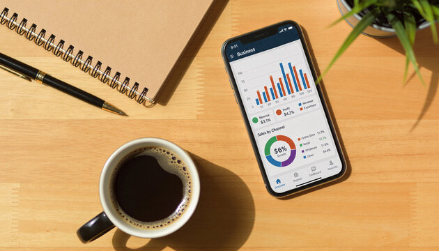 Smartphone displaying business analytics charts and sales data on a wooden office desk next to a coffee cup and notebook for financial planning and performance tracking