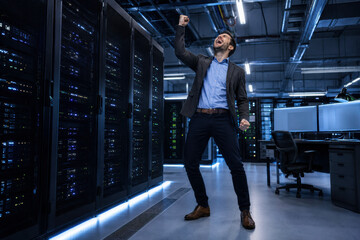 Obraz premium A businessman in a suit celebrates success with a joyful expression in a modern server room filled with data servers. Achievement and technology.