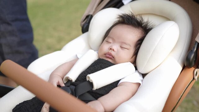 Soft white car seat holds sleeping baby. Crisp green grass surrounds the stroller. Baby's dark hair frames peaceful face. Mom's hand gently rests nearby. Calm, serene moment in open space