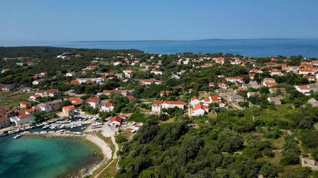 An aerial drone shot overlooking the charming, sun-drenched village on Silba Island, Croatia. The perspective captures traditional stone houses with bright red roofs nestled within lush green