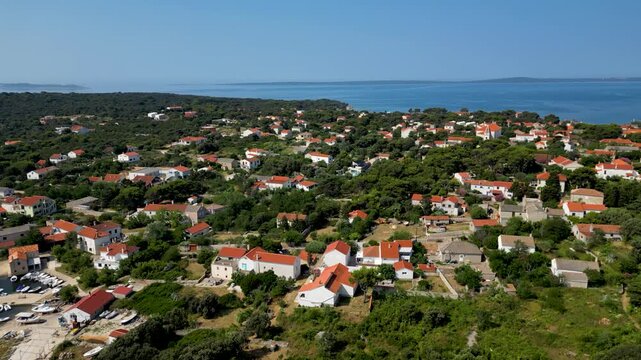 An aerial drone shot overlooking the charming, sun-drenched village on Silba Island, Croatia. The perspective captures traditional stone houses with bright red roofs nestled within lush green