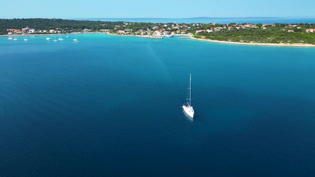 A stunning aerial drone perspective showcasing a white sailboat gliding through the crystal-clear, turquoise waters of Silba Island, Croatia. The shot beautifully captures the contrast between the