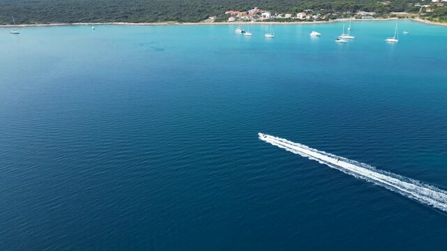 An exhilarating aerial drone shot over Silba Island, Croatia, capturing a water skier carving through the crystal-clear, turquoise sea behind a speeding motorboat. The top-down view beautifully frames