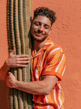 Smiling man hugging tall spiky cactus, enduring pain with adoring expression, humorous striking scene symbol of painful affection, thorny problem, summer adventure and seeking comfort in discomfort