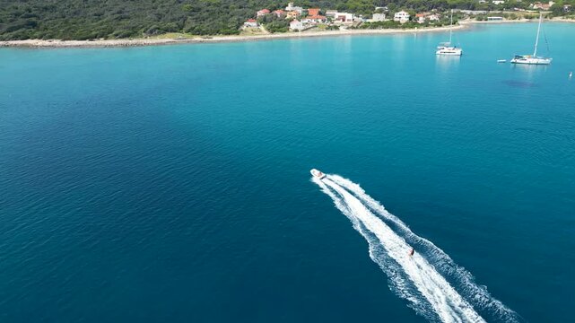An exhilarating aerial drone shot over Silba Island, Croatia, capturing a water skier carving through the crystal-clear, turquoise sea behind a speeding motorboat. The top-down view beautifully frames
