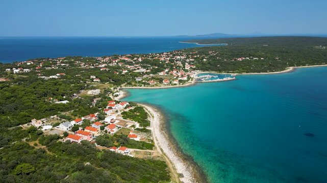 An aerial drone shot overlooking the charming, sun-drenched village on Silba Island, Croatia. The perspective captures traditional stone houses with bright red roofs nestled within lush green