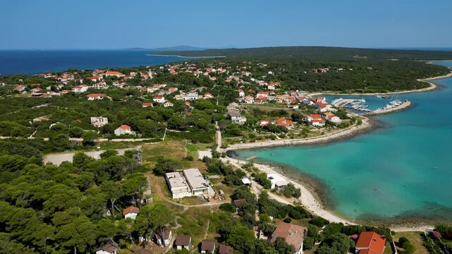 An aerial drone shot overlooking the charming, sun-drenched village on Silba Island, Croatia. The perspective captures traditional stone houses with bright red roofs nestled within lush green